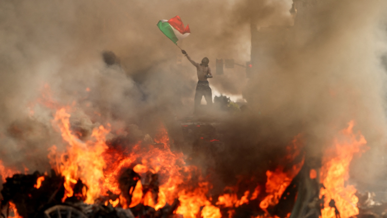 Un rivltoso sventola una bandiera messicana mentre fumo e fiamme si levano da un veicolo vicino al municipio di Los Angeles, 8 giugno 2025 (foto:  REUTERS/David Swanson).